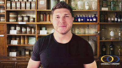 A man in a black shirt stands in front of vintage apothecary shelves filled with glass bottles and jars, representing natural health and wellness.